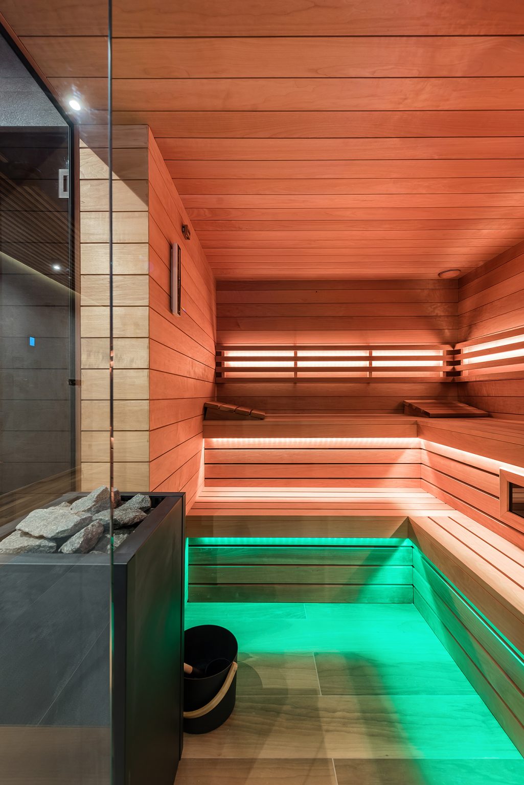Wnętrze nowoczesnej sauny z drewnianymi ławami, piecem z kamieniami i klimatycznym zielonym podświetleniem.