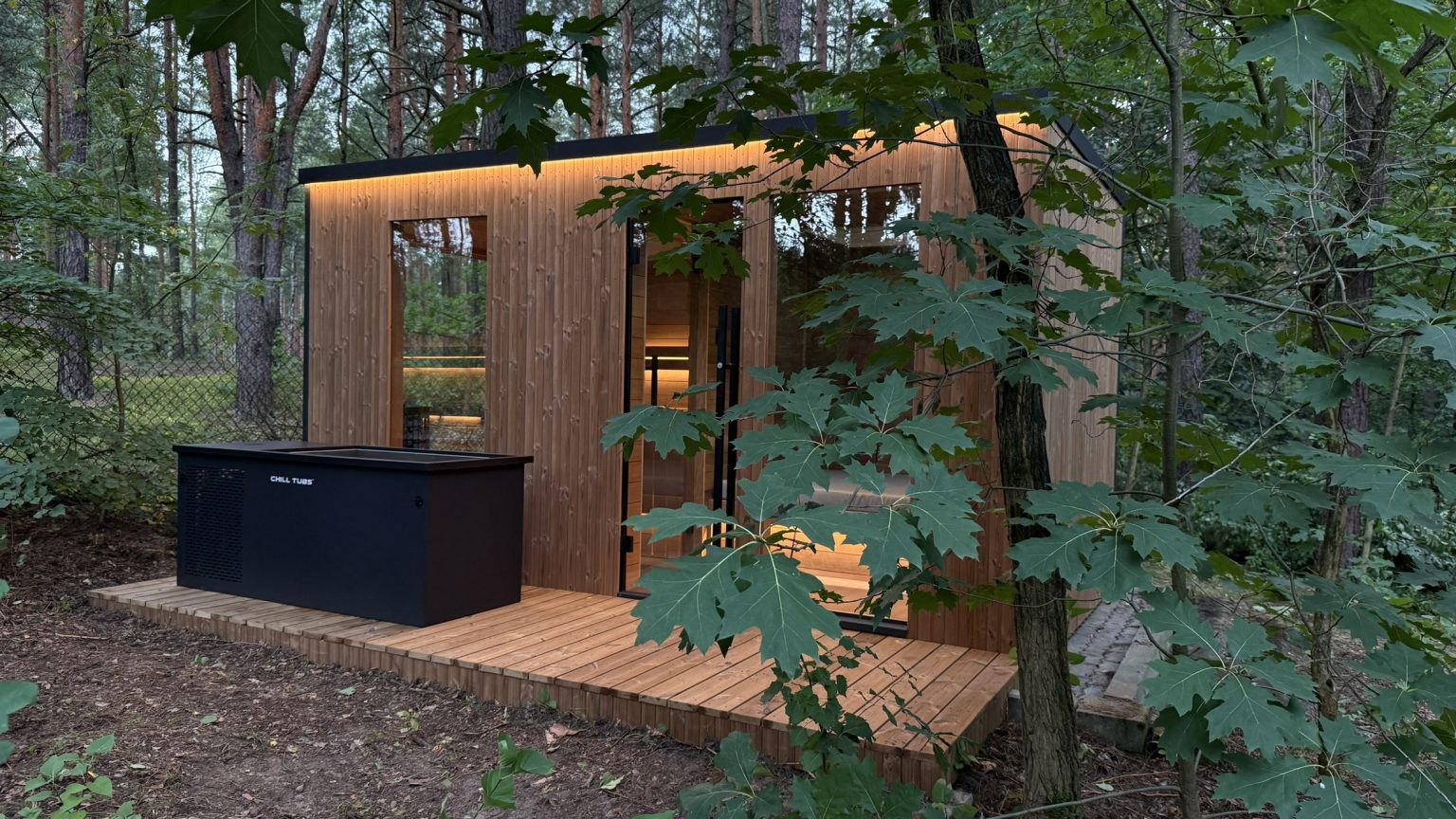 Nowoczesna sauna ogrodowa z bali z podświetlanym tarasem i schładzalnikiem Chill Tubs w leśnym otoczeniu.