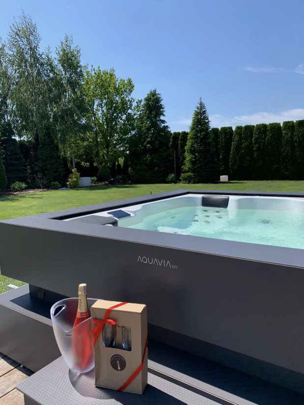 Wanna ogrodowa Aquavia Spa z butelką szampana i kieliszkami w ozdobnym pudełku na tarasie w ogrodzie.