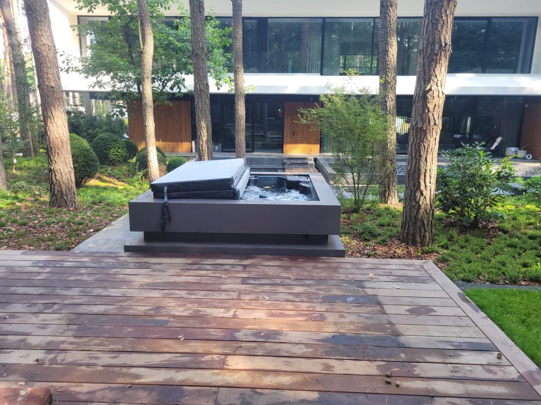 Zewnętrzne jacuzzi z częściowo otwartą pokrywą na drewnianym tarasie w ogrodzie przy nowoczesnym domu.