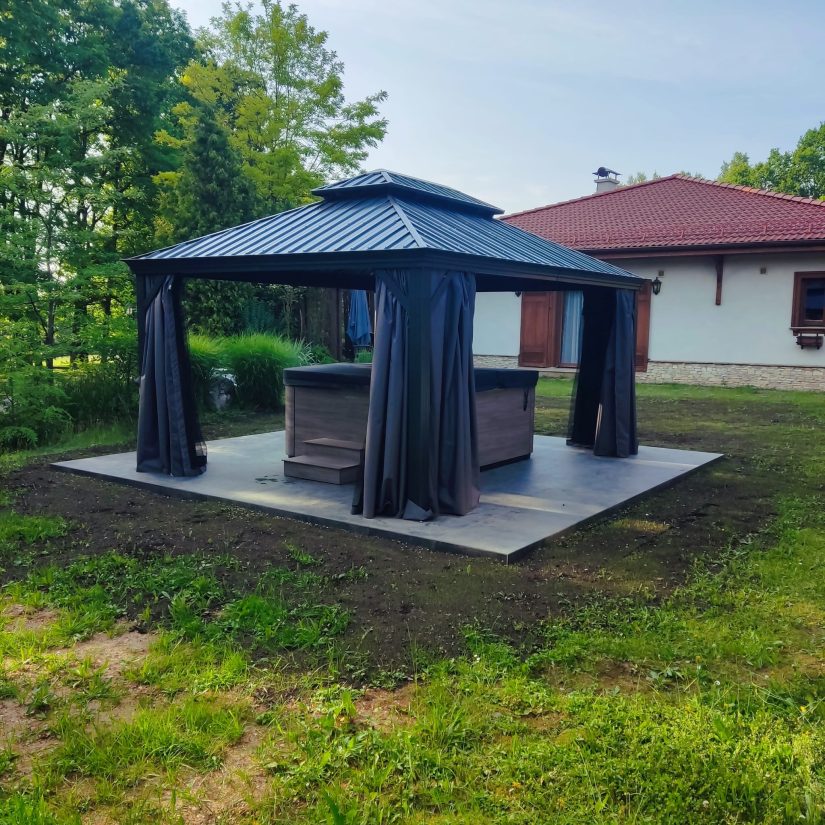 Wanna ogrodowa zadaszona pawilonem na betonowej platformie w ogrodzie.