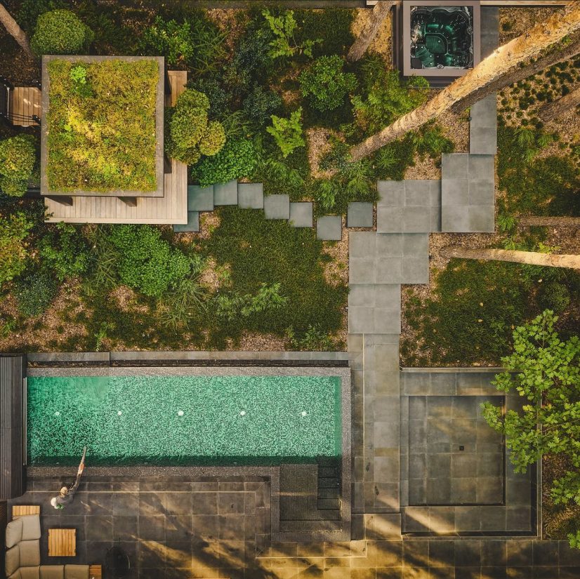 Widok z góry na nowoczesny ogród z basenem, jacuzzi, ścieżkami z płyt kamiennych i altaną z zielonym dachem.