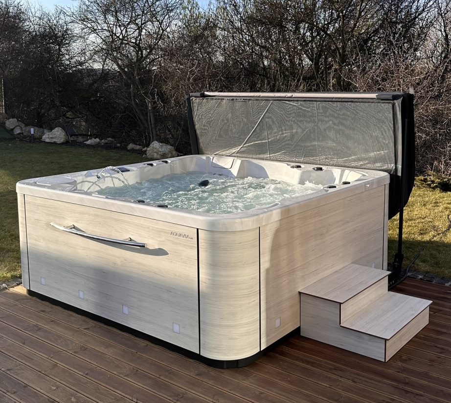 Wanna z hydromasażem Aquavia Spa z podniesioną pokrywą na drewnianym tarasie w ogrodzie.