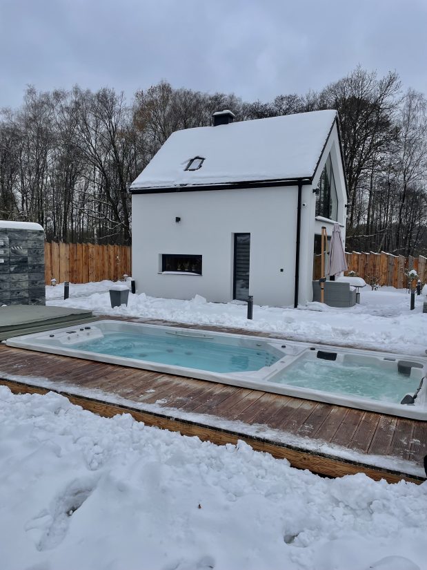 Biały dom z zaśnieżonym dachem i nowoczesne jacuzzi na drewnianym tarasie w zimowym ogrodzie.
