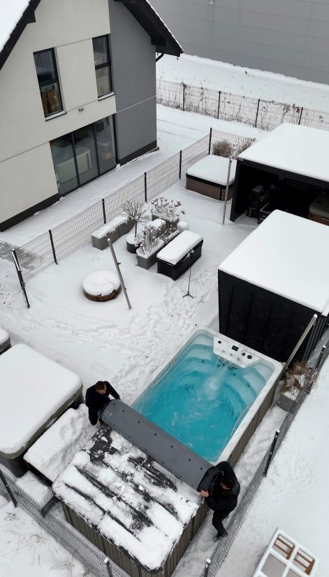 Dwóch mężczyzn odsłania przykrycie ogrodowego basenu z hydromasażem w zimowy dzień.