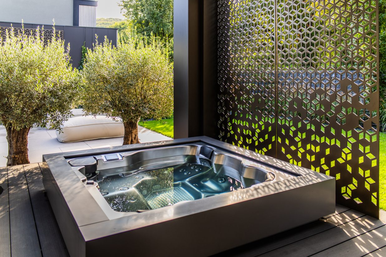 Zewnętrzne jacuzzi na tarasie obok ozdobnego ażurowego panelu i drzewek oliwnych.