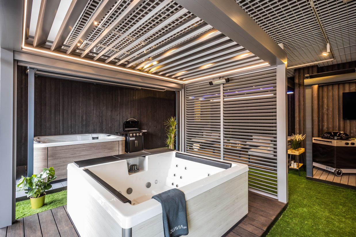 Nowoczesne jacuzzi Quavia Spa ustawione na tarasie z pergolą, roślinami i grillem.