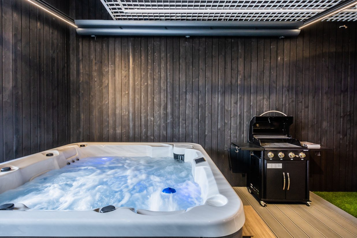 Wanna z hydromasażem i grill gazowy na tarasie z ciemnym drewnianym wykończeniem ścian.