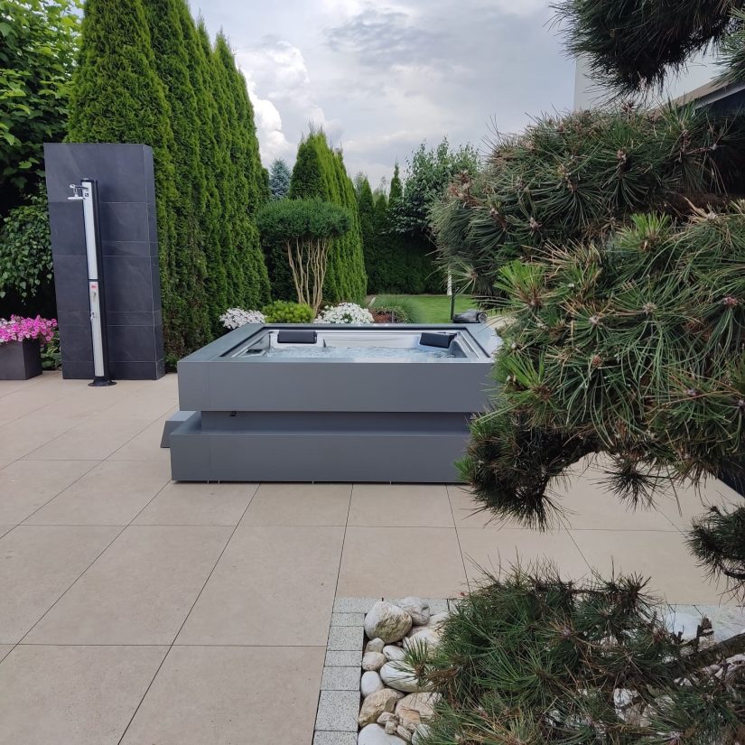 Nowoczesne jacuzzi ogrodowe na tarasie z zewnętrznym prysznicem w otoczeniu iglaków.